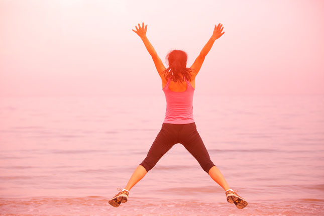 Permalink to Improve Health and Wellbeing and Enjoy a Holiday Too Woman jumping into the air by the beach